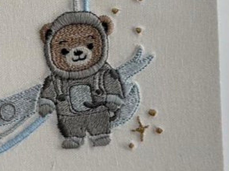 Ursinho vestido de astronauta, com capacete arredondado, traje cinza e mochila, sentado sobre a faixa