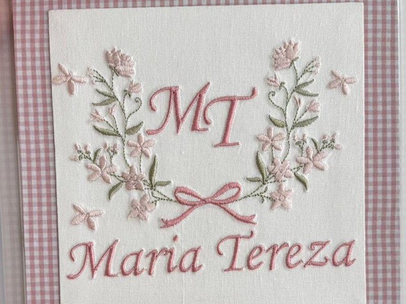 'MT' em rosa dentro de uma coroa de flores e folhas com la�o na base, borboletas em volta e 'Maria Tereza' bordado abaixo