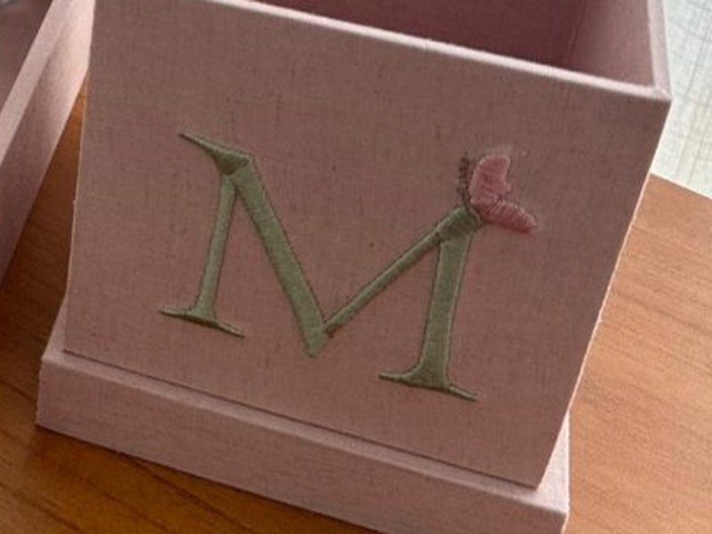 O cachep� quadrado rosa com o 'M' verde escuro bordado e uma florzinha rosa ao lado da letra
