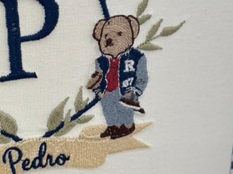 O ursinho de jaqueta com a letra R segurando uma raquete, bordado � direita do escudo