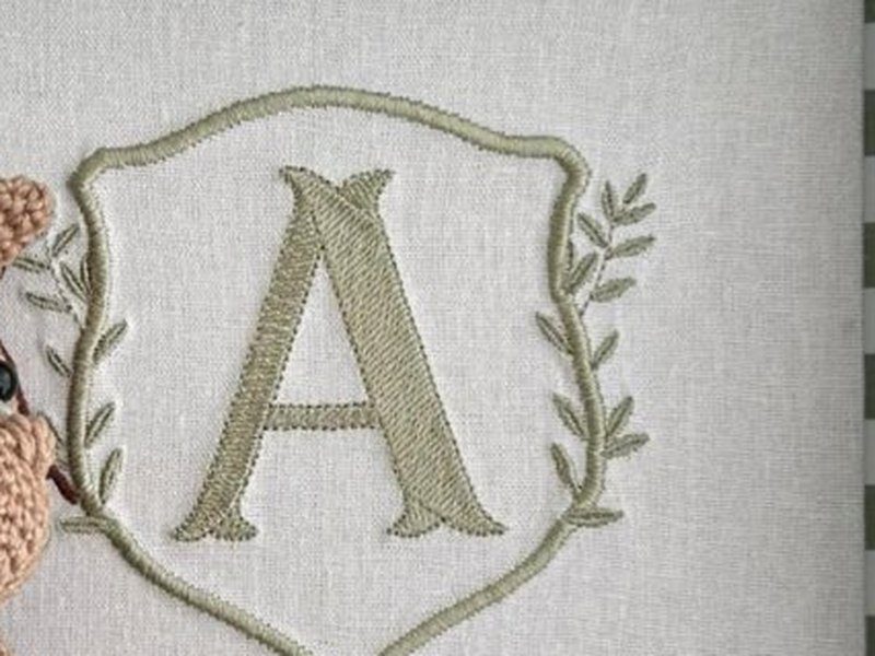 Escudo de contorno verde com a letra A ao centro e um ramo de folhas � direita, tudo bordado em verde sobre linho