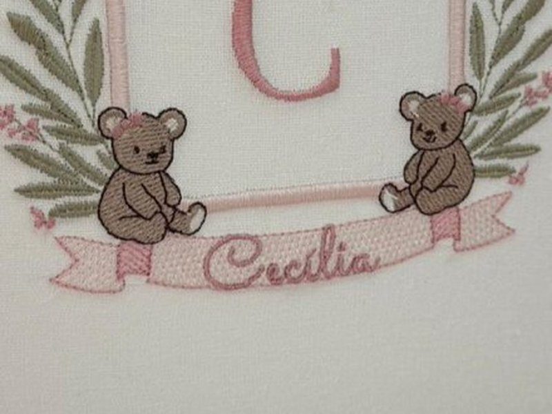 Os dois ursinhos bordados sentados, um de cada lado, apoiados sobre a faixa rosa com 'Cec�lia'