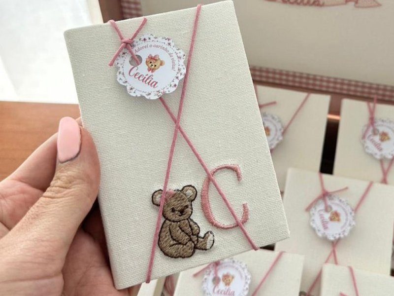 Capa de linho cru com um ursinho marrom bordado e a letra C em rosa, cord�o rosa cruzado e tag rendada