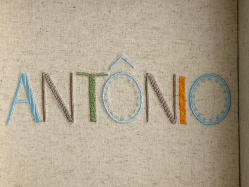 O nome ANT�NIO bordado por inteiro em letra bast�o, cada letra numa cor e num tipo de ponto diferente, com pontinhos em volta do O final