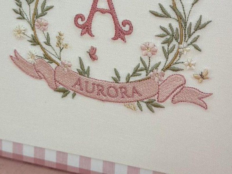 Faixa rosa bordada com AURORA em caixa alta e folhinhas verdes nas pontas