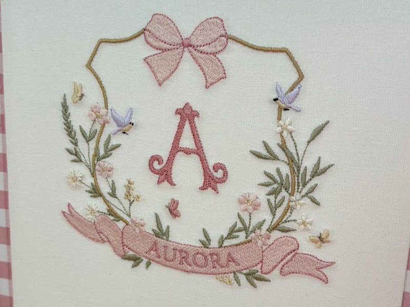 Escudo com contorno em fio dourado, la�o rosa bordado no topo, A ornamentada ao centro e ramos floridos com passarinhos lil�s dos dois lados