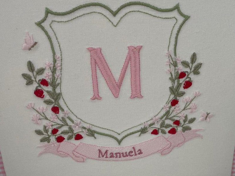 Escudo contornado em verde com M rosa ao centro e ramos carregados de morangos vermelhos, florzinhas rosa e brancas e borboletas