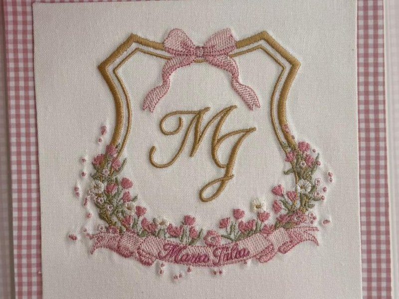 Bras�o com contorno em fio dourado, la�o rosa no topo, MJ em cursiva dourada e guirlanda de flores rosa em relevo
