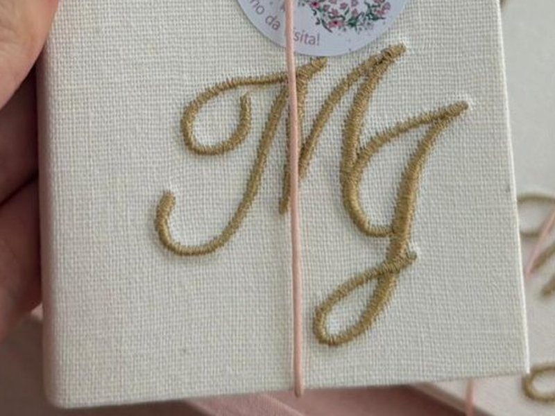 Livrinho em linho cru com MJ bordado em fio dourado grande, atravessado por el�stico rosa
