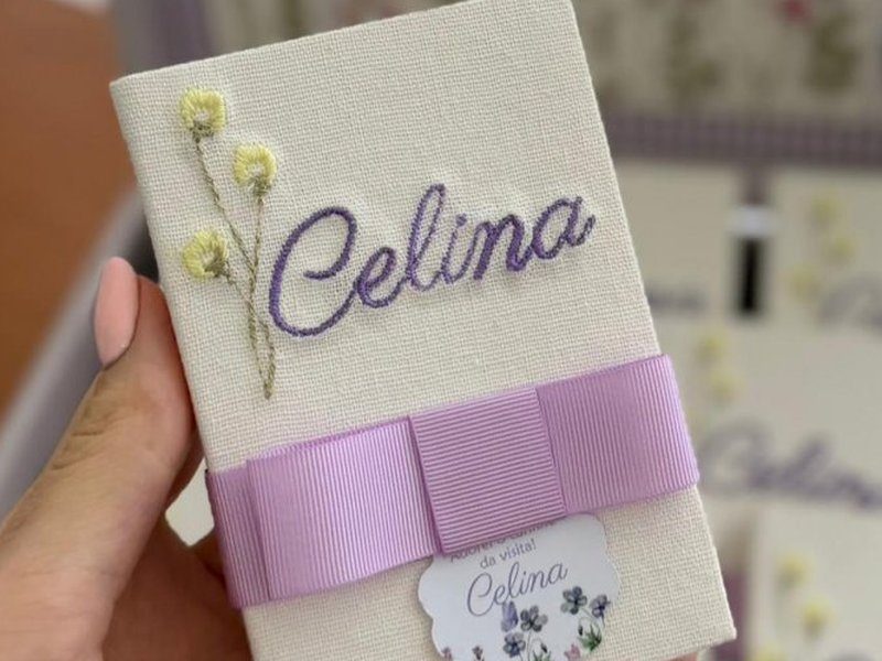 Livrinho com Celina em cursiva roxa e ramo de tr�s flores amarelas, cintado por la�o largo lil�s com tag redonda