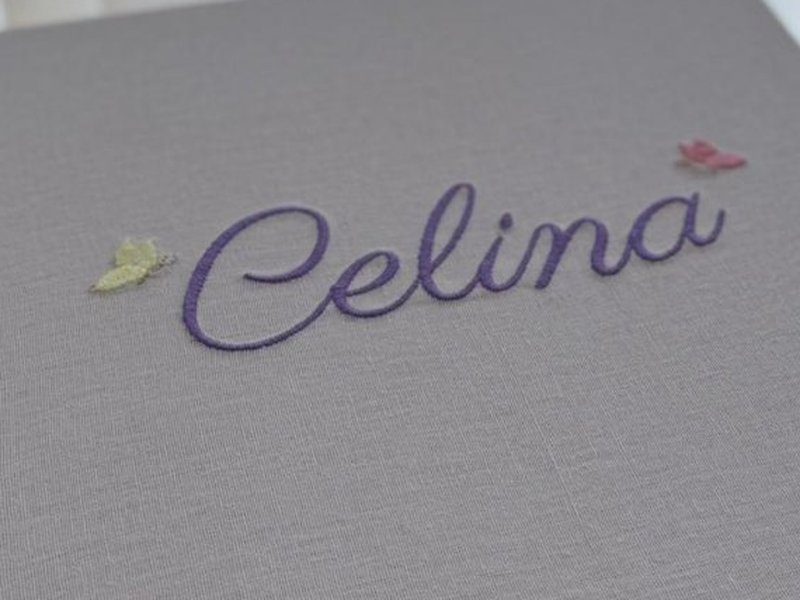 Celina bordado em roxo sobre o linho cinza claro da tampa fechada, entre uma borboleta amarela e uma rosa min�sculas