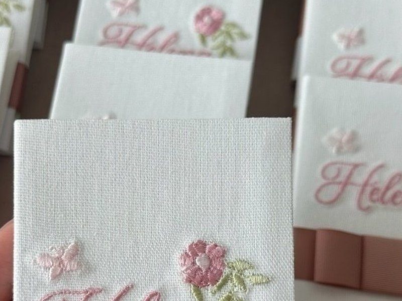 Flor rosa bordada em relevo alto com folhas verdes e borboleta ao lado do nome Liz em cursiva ros�