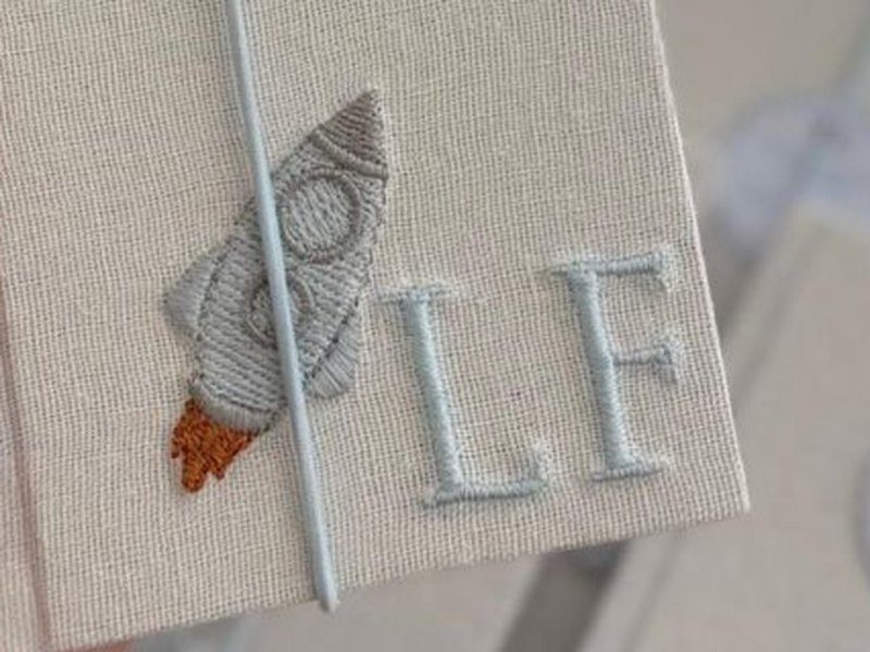 Caixinha-livro com um foguete bordado em cinza e propuls�o laranja ao lado das iniciais LF em azul-claro