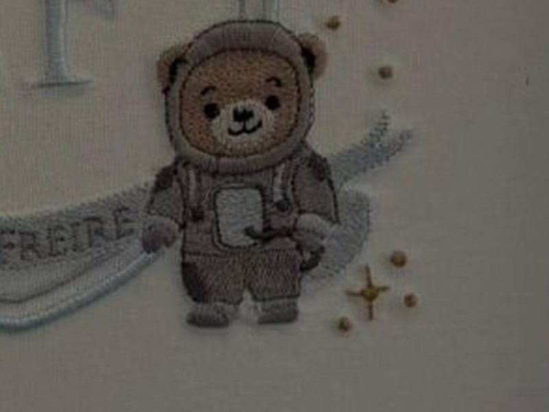 Urso bordado vestido de traje de astronauta com capacete, ao lado do bras�o