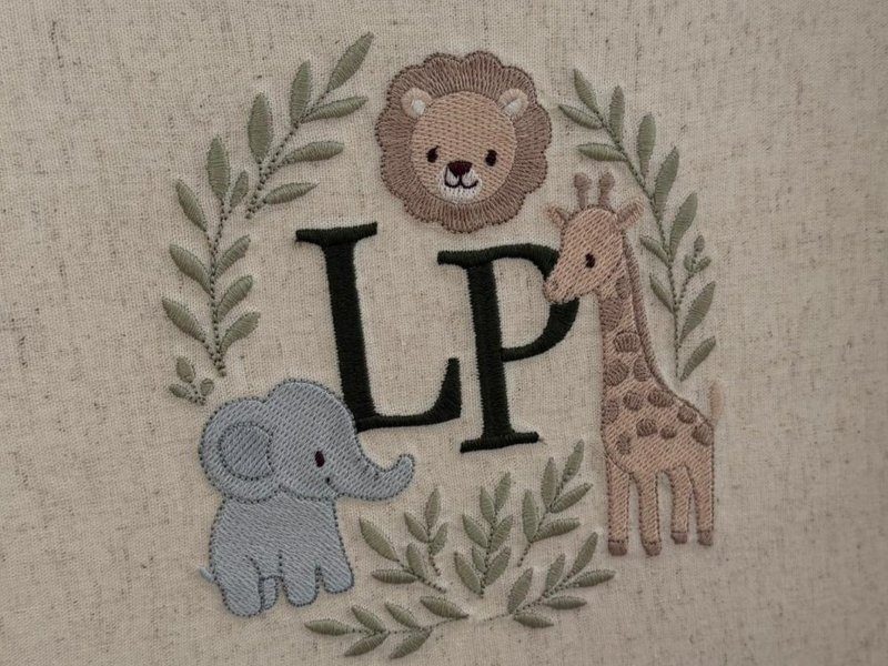 Bras�o bordado na tampa: iniciais LP em verde escuro entre le�o, elefante e girafa, dentro de coroa de ramos de oliveira sobre linho cru