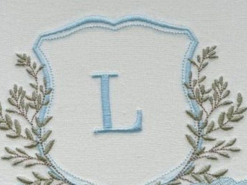 Escudo de contorno azul-claro com a letra L azul ao centro e coroa de folhas verde escuro em volta
