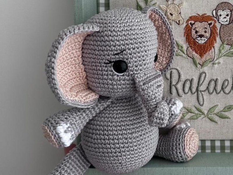 Elefante de croch� cinza com orelhas e patas rosa-claro, tromba curvada e olhos pretos, sentado na frente do quadro