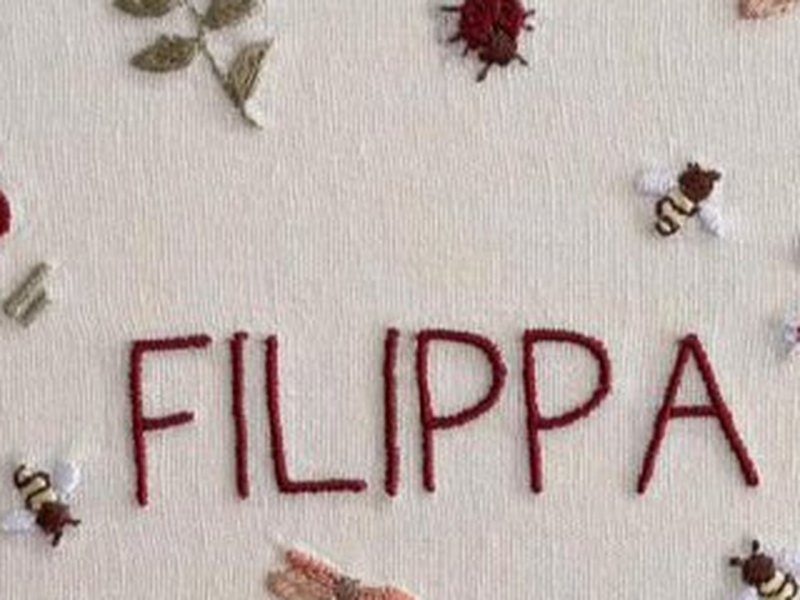 O nome FILIPPA bordado por inteiro em ponto cheio vermelho escuro, com joaninhas e abelhas ao redor