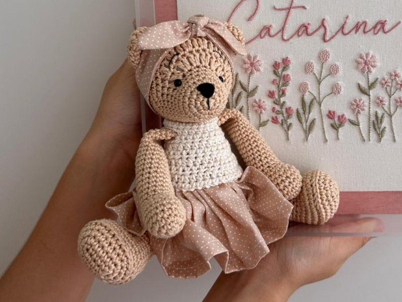 Ursa de croch� bege com vestido de tecido bege de po� branco, gola branca de croch� e la�o de tecido amarrado na cabe�a