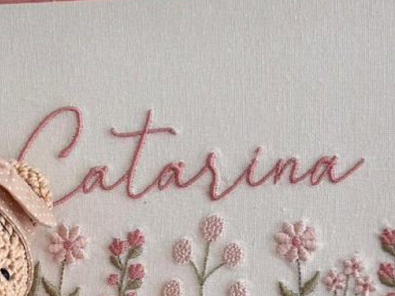O nome Catarina bordado por inteiro em cursiva rosa, acima da faixa de flores