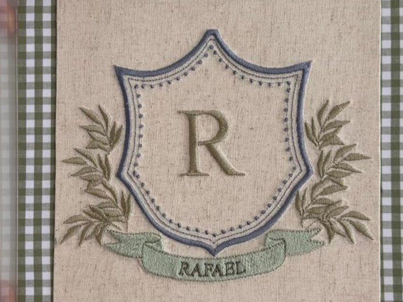 Escudo bordado contornado em azul com pontilhado, R em bege ao centro e ramos de folhas verde escuro dos dois lados