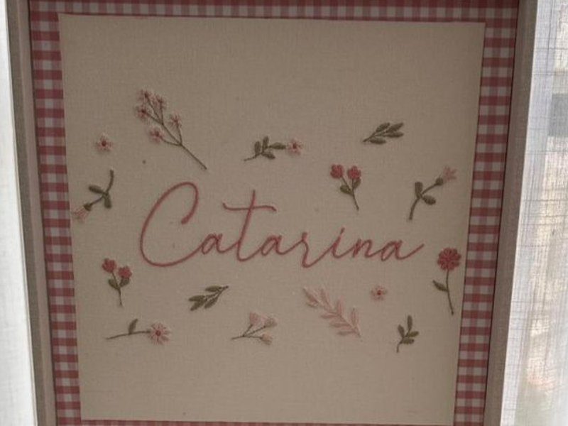 Catarina em cursiva rosa ao centro, cercado por ramos, folhas verdes e florzinhas rosa soltas pelo painel