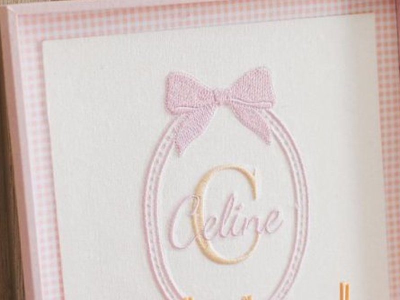 Monograma C em fio dourado dentro de moldura oval rosa com o nome Celine em cursiva escrito por cima da letra, e o la�o bordado acima