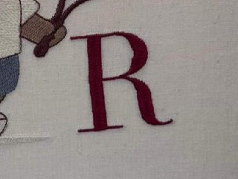 A letra R serifada bordada em vinho ao lado do urso