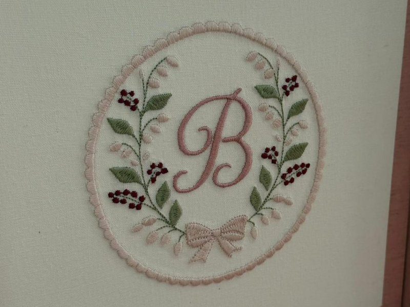 Monograma B em cursiva ros� dentro de guirlanda oval de borda em picot, com folhas verdes, bot�es claros, frutinhas vinho e la�o na base