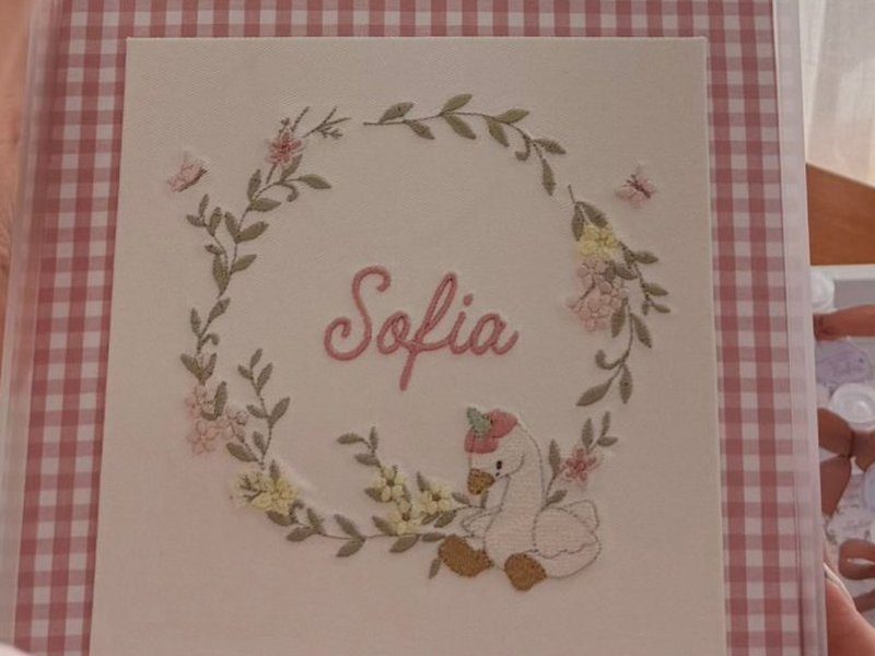 Guirlanda circular de folhas verdes com flores rosa e amarelas e borboletas, o nome Sofia em cursiva rosa ao centro e um gansinho de toquinha bordado na base