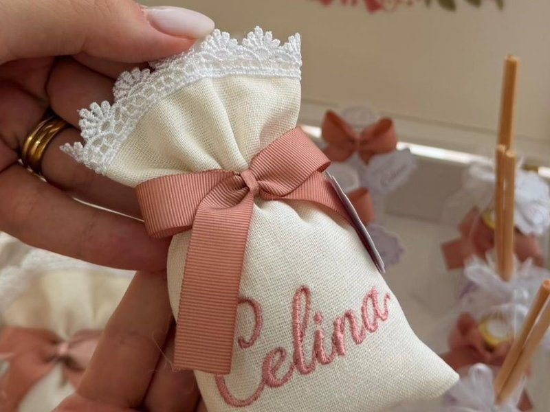Sach� de linho com barrado de renda branca, la�o tom tijolo e o nome Celina bordado em rosa