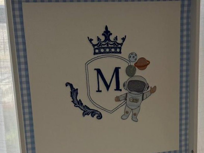 Escudo bordado com a letra M, coroa acima, astronauta ao lado direito segurando planeta e lua, e ramo de folhagem a esquerda