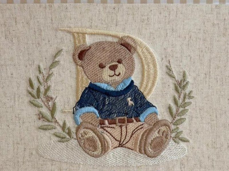 Urso bordado de su�ter azul escuro e bermuda clara sentado sobre um carrinho, com a letra D bege atr�s dele e ramos de oliveira dos dois lados