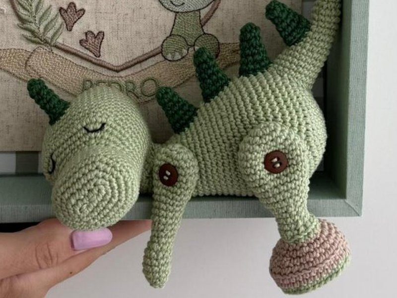 O dinossauro de croch� verde com espinhos escuros nas costas e olhos de bot�o, apoiado na borda do quadro