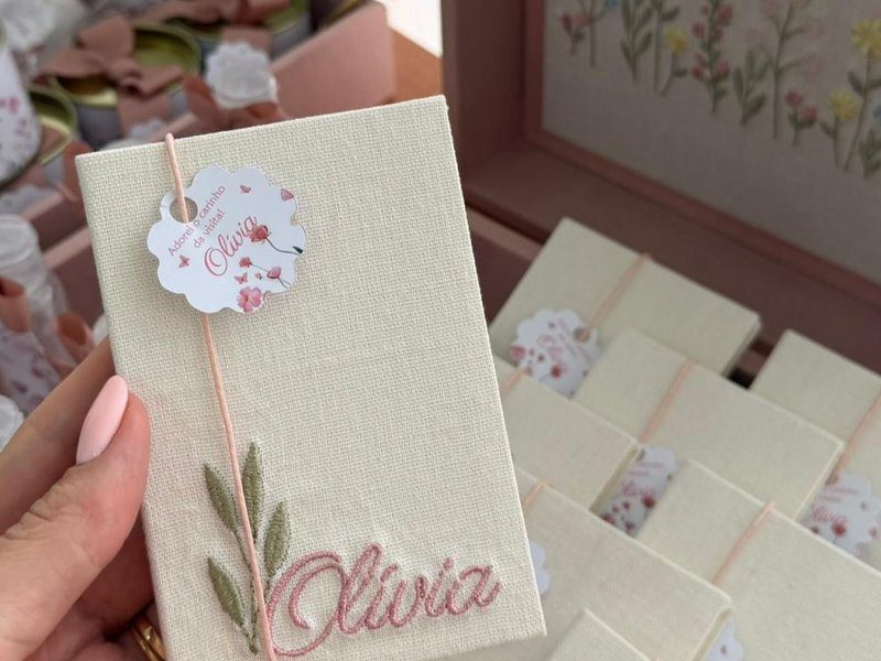 Livrinho de capa de linho cru com o nome Olivia bordado em rose e um ramo de folhas verdes, cordao rose e tag redonda