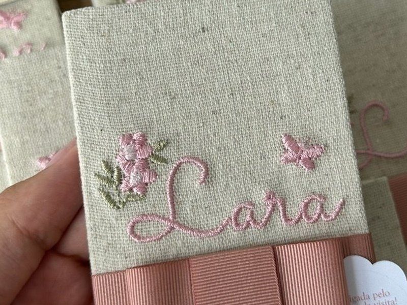 Capa de linho com o nome Helena bordado em rose, uma flor rosa de miolo em relevo, uma borboleta e uma haste verde, sobre cinta de gorgur�o rose