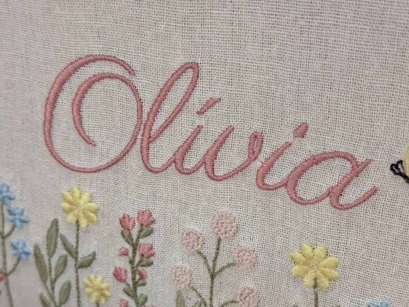 O nome Ol�via em cursiva ros� sobre um campo de flores bordado - margaridas amarelas, mios�tis azuis, bot�es rosa em ponto cheio e caules verdes - com uma borboleta amarela de antenas pretas
