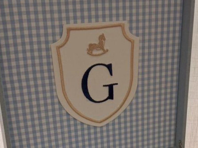 Escudo bordado com contorno bege, cavalinho de balan�o no topo e a inicial G em azul escuro, sobre fundo xadrez