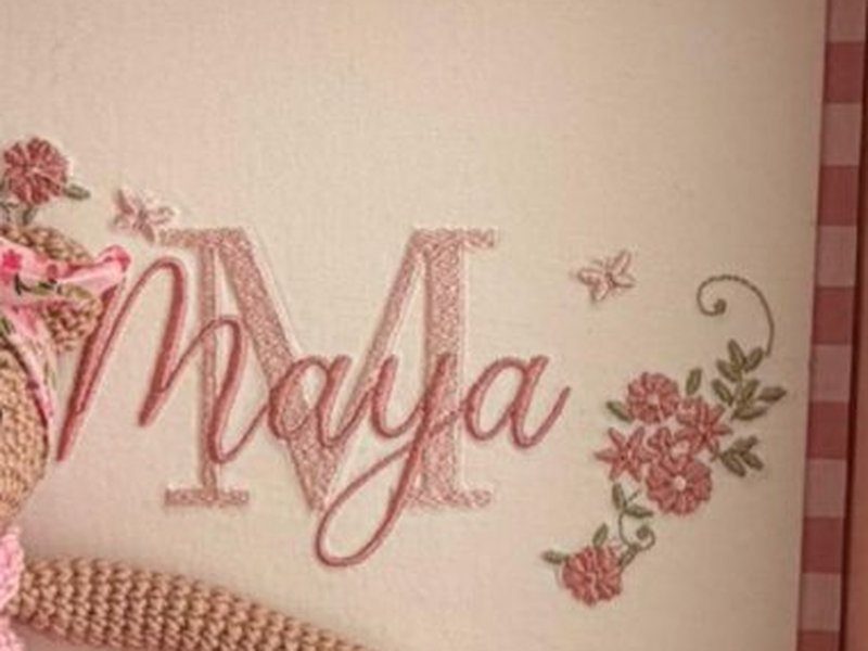 A letra M bordada em rosa com o nome Maya sobreposto em cursiva, ao lado do ramo de rosas