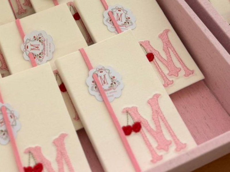 Capa creme do livrinho com a inicial M bordada em rosa e um par de cerejas vermelhas, atravessada pelo el�stico