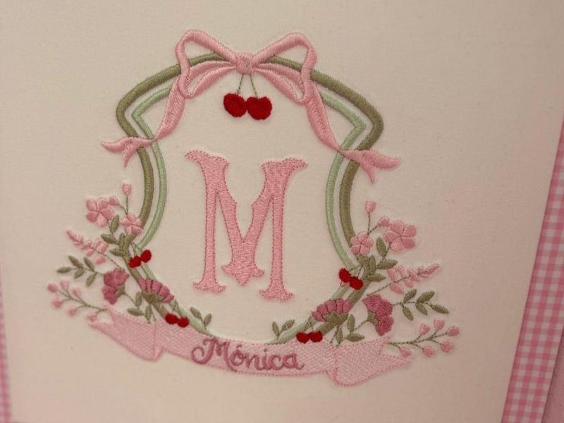 Bras�o bordado: la�o rosa, par de cerejas vermelhas, moldura oval verde, inicial M rosa e faixa com M�nica