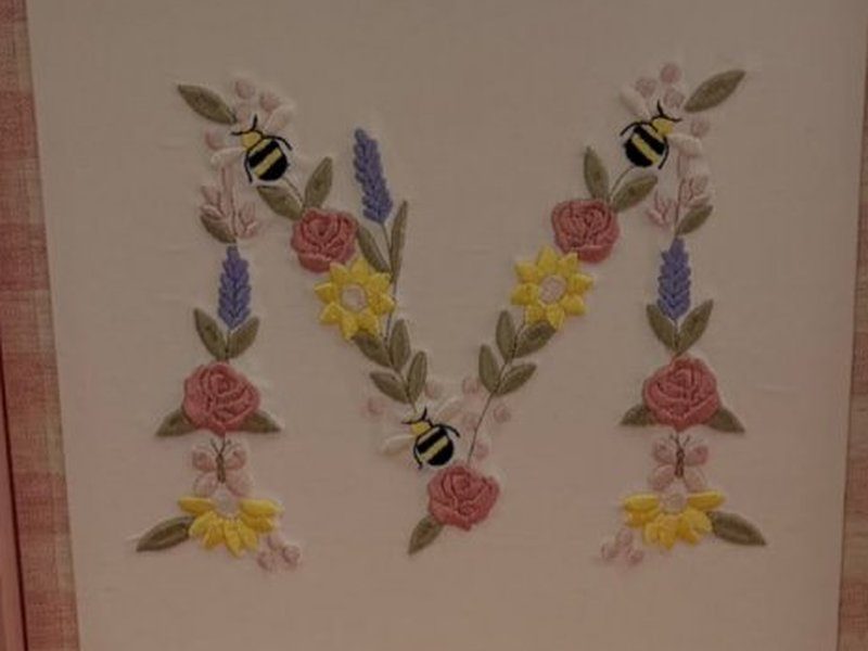 A inicial M desenhada inteiramente com flores bordadas - rosas, margaridas amarelas, lavandas - e tr�s abelhas pousadas nela