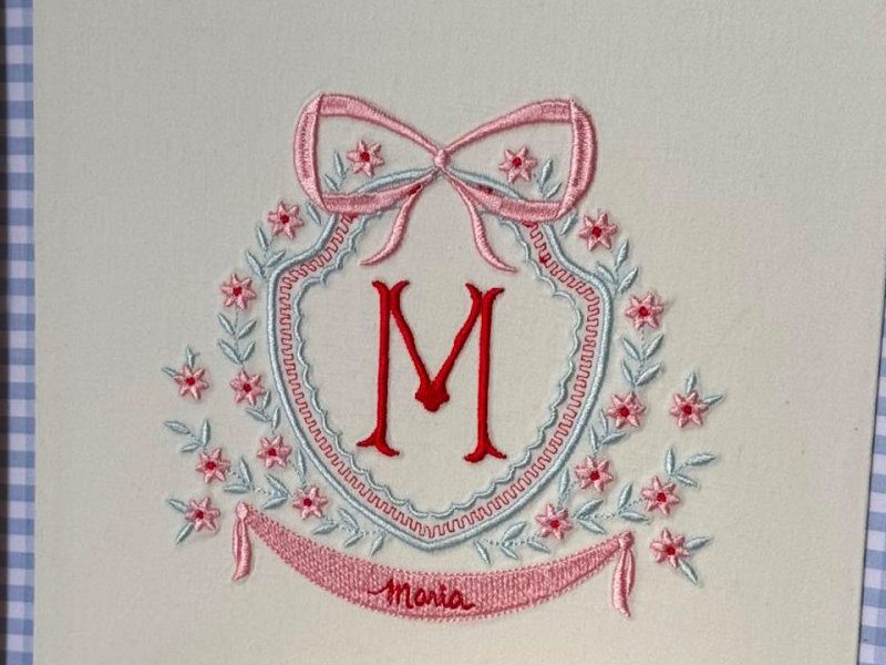 Bras�o com la�o rosa, moldura oval azul-clara rendilhada, flores rosa, inicial M em vermelho e faixa rosa com Maria