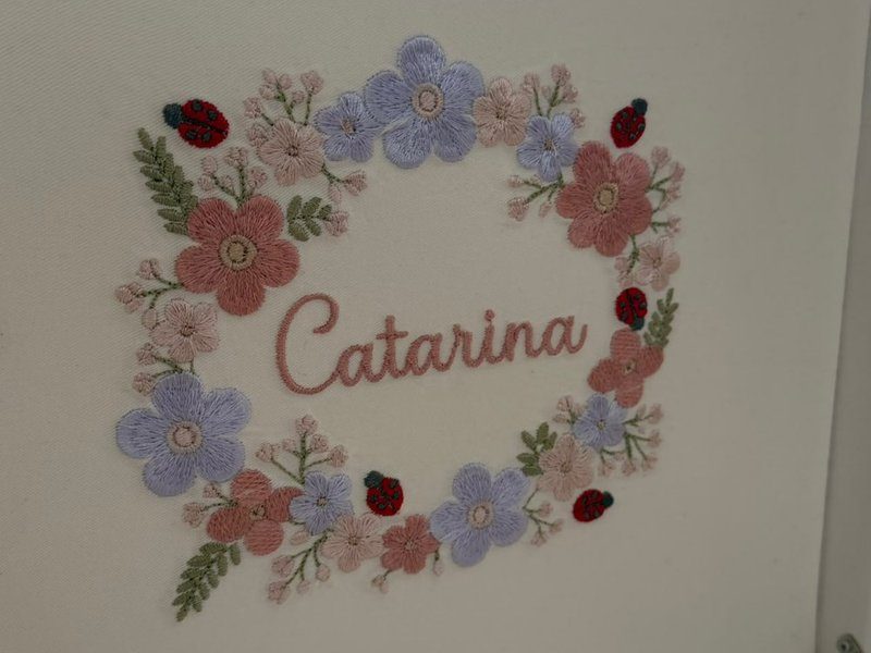 Coroa de flores bordadas em rosa, lil�s e cru, com cinco joaninhas vermelhas de cabe�a preta, e Catarina em cursiva ao centro