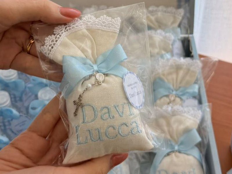 Sach� de linho com renda guipir na boca, la�o azul com um tercinho de contas e crucifixo prateado amarrado, e o nome Davi Lucca bordado em duas linhas