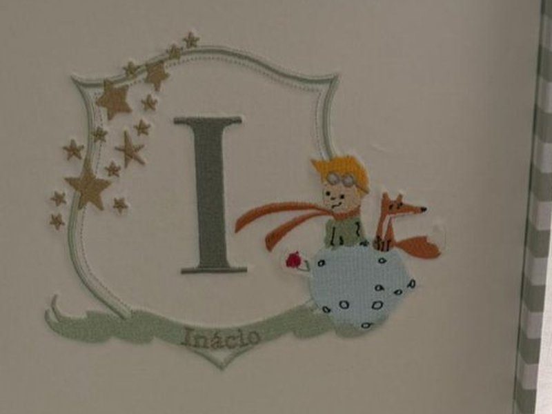 Escudo bordado com a inicial I, estrelas douradas, o Pequeno Pr�ncipe de capa laranja sobre o asteroide, a raposa e a rosa vermelha, e faixa com In�cio