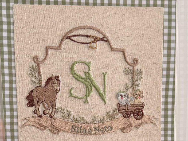 Bras�o bordado no linho: cavalo � esquerda, monograma SN em verde ao centro, carro�a com dois carneirinhos � direita, chicote com estribo no alto e faixa com Silas Neto