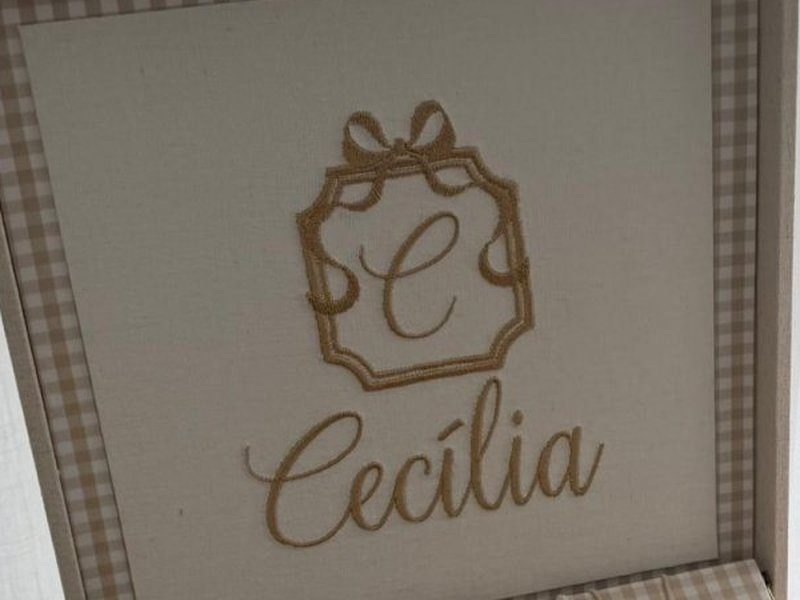 Bras�o bordado em fio dourado: moldura recortada com la�o no topo, a inicial C ao centro e Cec�lia em cursiva abaixo