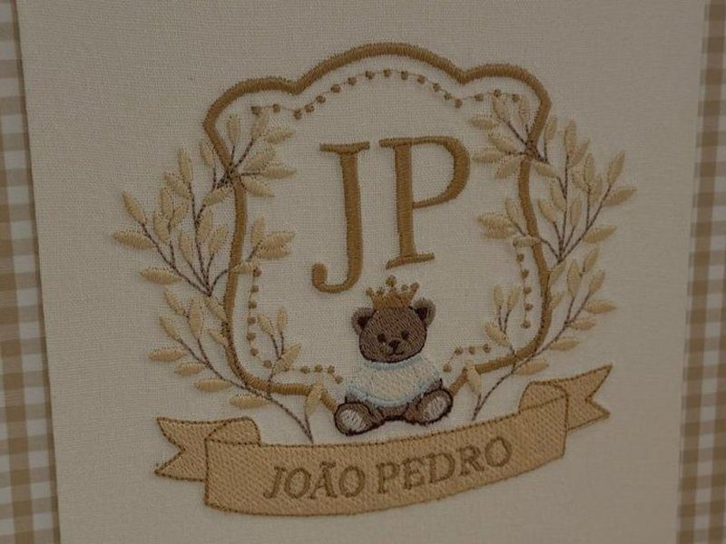 Escudo bordado em fio dourado com JP ao centro, ramos de louro com ponto de bolinha, ursinho de su�ter azul com coroa dourada e faixa bordada com JO�O PEDRO