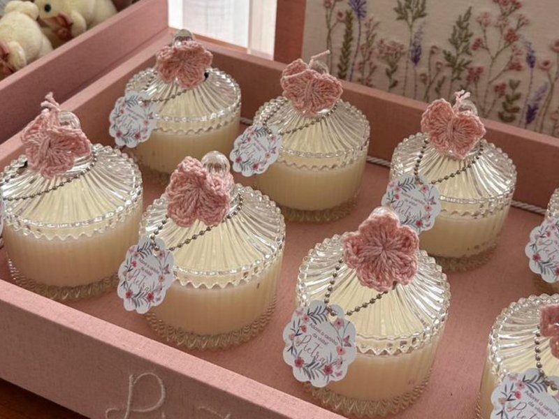 Velas em pote de vidro canelado com tampa, cada uma com uma flor de croch� ros� no topo e tag redonda com o nome